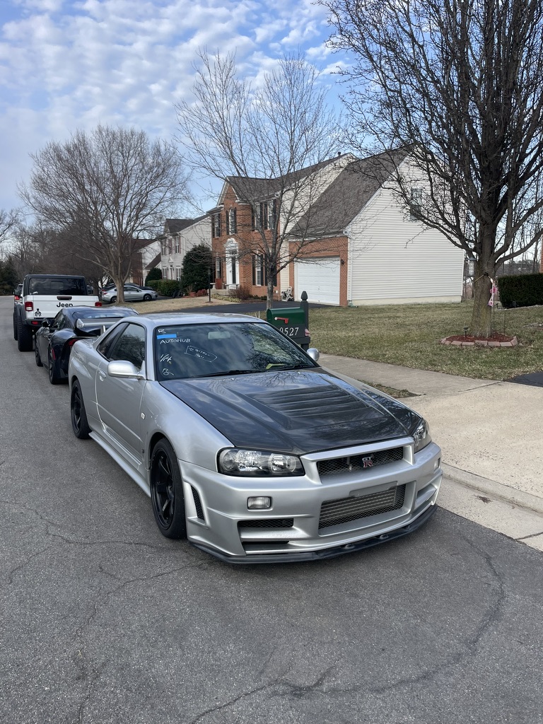 Silver Nissan Skyline GT-R R34 V-Spec with a carbon-fibre bonnet, front three-quarter view, parked on a residential street on black aftermarket wheels.